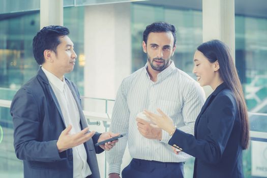 Two handsome young businessmen and lady in classic suits are holding cups of coffee, talking and smiling, standing outside the office building.