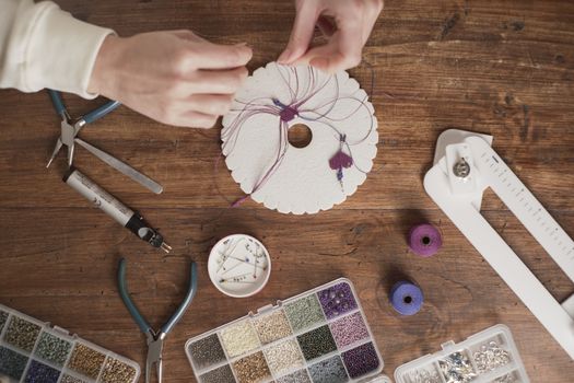 Lifestyle concept, work from home to reinvent your life: top view of woman hands making macrame knots the fuchsia thread creating an earring on kumihimo tools on wooden table