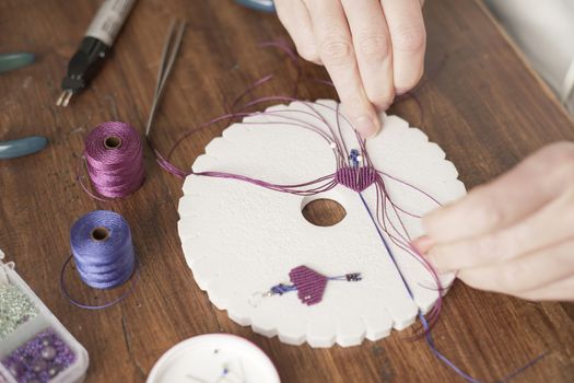 Lifestyle concept, work from home to reinvent your life: close-up of woman hands making macrame knots the fuchsia thread creating an earring on kumihimo tools on wooden table