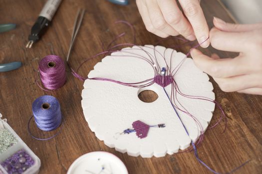 Lifestyle concept, work from home to reinvent your life: close-up of woman hands making macrame knots the fuchsia thread creating an earring on kumihimo tools on wooden table