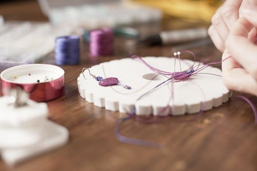 Lifestyle concept, work from home to reinvent your life: close-up of woman hands making macrame knots the fuchsia thread creating an earring on kumihimo tools on wooden table