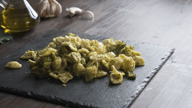 Closeup of a slate plate with steaming orecchiette with broccoli, typical Apulian recipe, in backlight on dark wooden table with olive oil and garlic on background in bokeh effect