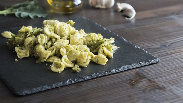Closeup of a slate plate with steaming orecchiette with broccoli, typical Apulian recipe, in backlight on dark wooden table with olive oil and garlic on background in bokeh effect