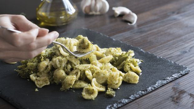 Closeup of a woman's hand that pick up with a fork steaming orecchiette with broccoli, typical Apulian recipe, in backlight on dark wooden table with olive oil and garlic on background in bokeh effect