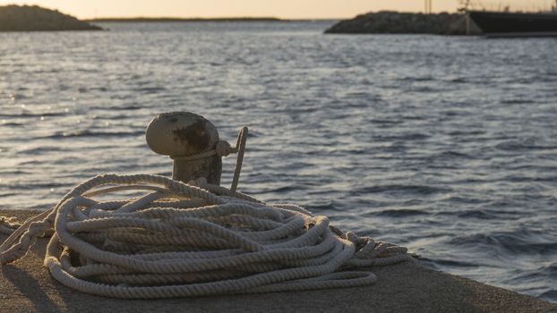 Backlight foreground at sunrise or sunset of a mooring bollard with the rope tied and resting on the ground and various boats in the background in back light