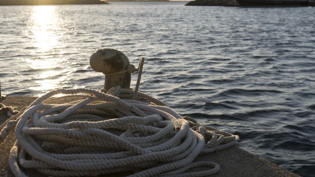 Backlight foreground at sunrise or sunset of a mooring bollard with the rope tied and resting on the ground and various boats in the background in back light