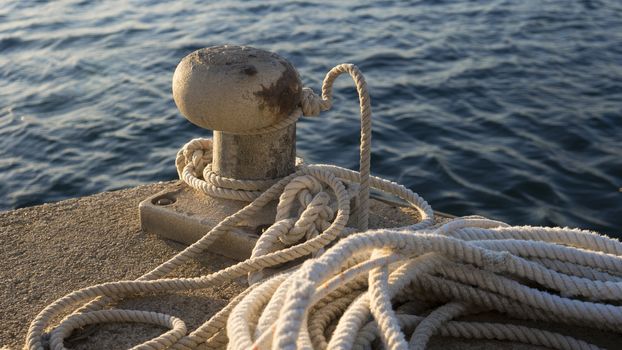 Backlight foreground at sunrise or sunset of a mooring bollard with the rope tied and resting on the ground and various boats in the background in back light