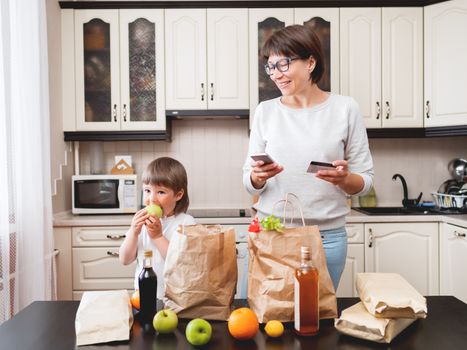 Woman and toddler boys sorts out purchases in the kitchen. Kid bites an apple. Grocery delivery in paper bags. Subscription service from grocery store. Mother and son at kitchen.
