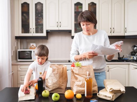 Woman and toddler boys sorts out purchases in the kitchen. Grocery delivery in paper bags. Subscription service from grocery store. Mother and son at kitchen.