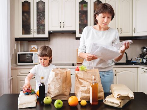 Woman and toddler boys sorts out purchases in the kitchen. Grocery delivery in paper bags. Subscription service from grocery store. Mother and son at kitchen.