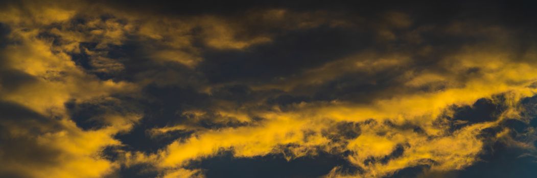 Panorama golden fluffy clouds illuminated by disappearing rays at sunset and dark cumulonimbus floating across blue sky to change season weather. Soft focus, motion blur sky landscape.