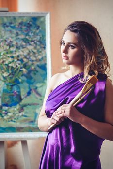 model girl in a blue dress in an art workshop with brushes, a palette and paintings