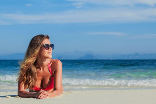 Glamorous long haired young woman in bikini and sunglasses lying on tropical beach, copy space for content