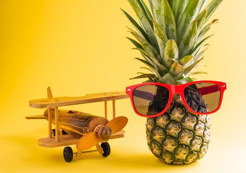 Celebrate Summer Pineapple Day Concept, funny fresh pineapple in sunglasses stands with a model plane, studio shot isolated on yellow background, Holiday summertime in tropical