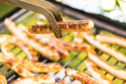 Grilled sausages on barbecue grill with close-up of fried bratwurst holding with barbecue tongs