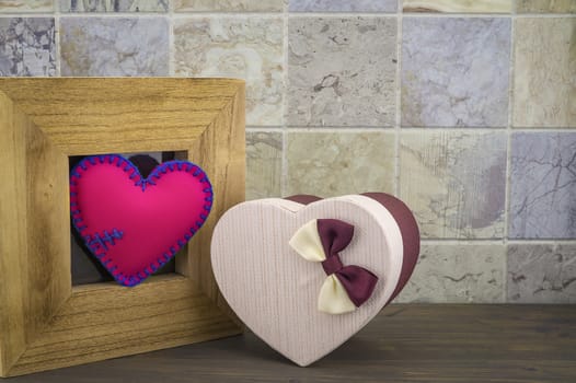 Handcrafted pink heart in a wooden frame with a heart shaped gift box against a tiled background