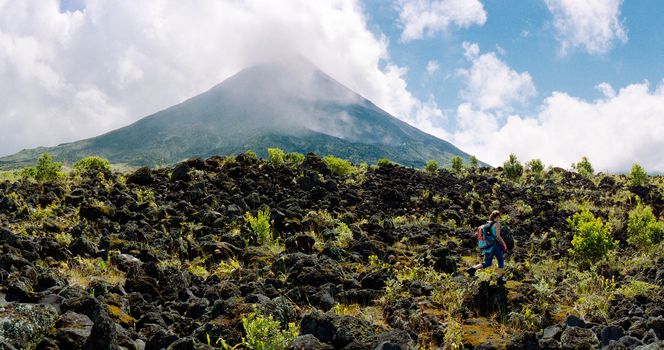 wide panoramic view on the volcano arenal in a national park in Costa rica withs some clouds. A women is walking in the foreground on lava rocks with some foliage between them