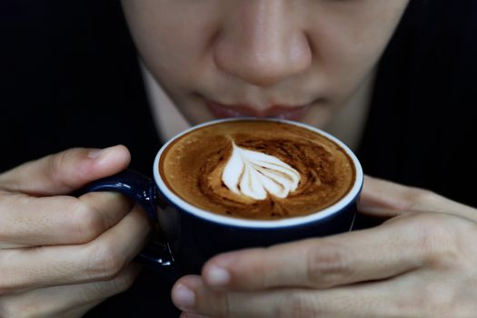Close up of hot cappuccino coffee in the cafe, Heart shape cream coffee in blue cup. Hands women holding hot coffee in cup Asian woman smelling and drinking hot coffee with feeling good in coffee shop