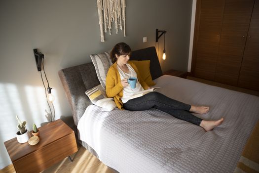 Beautiful woman on bed with a cup of coffee and reading a book