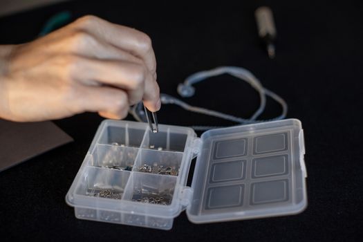 Lifestyle concept, work from home to reinvent your life: close-up of woman hands making macrame knotted jewelry picks up a bead with tweezers from the organizer box and tools on black work table