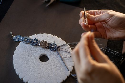 Lifestyle concept, reinvent your life and your job: close-up detail of woman hands making macrame knotted jewel with the fingers threading the bead into the nylon thread on black work table