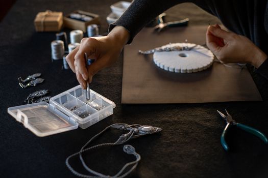 Lifestyle concept, work from home to reinvent your life: close-up of woman hands making macrame knotted jewelry picks up a bead with tweezers from the organizer box and tools on black work table