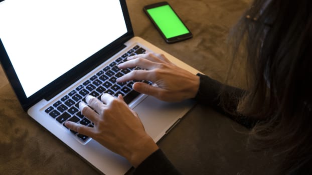 Couple relationships and dating in modern times concept: closeup of woman's hands typing on laptop keyboard with blank white screen in low light with smartphone alongside with green blank screen