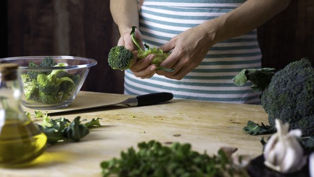 Moments of everyday life in Mediterranean cuisine: young female cook cleans broccoli on a wooden chopping board with extra virgin olive oil, garlic, pepper and knife on top