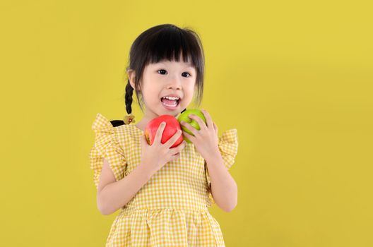 happy little asian girl holding green and red apple isolated on yellow background.