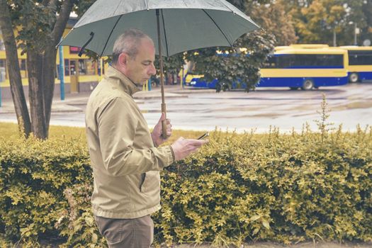 A man standing at the bus station with an umbrella waiting for a bus. Man waiting on a bus, texting on mobile phone in a rainy day. Rainy day in a local town.