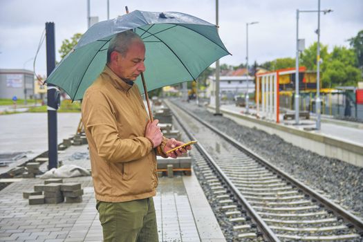 Portrait of a mature man texting on smartphone with an umbrella standing on the platform of a train station. Unfinished railway line. When the train arrives.. Funny concept of vain situation.