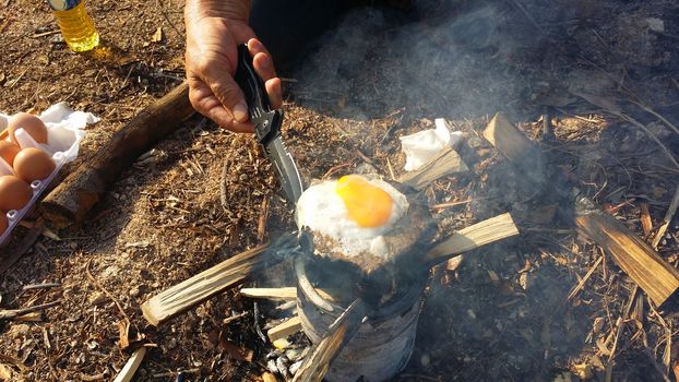 person cooking fried eggs in nature camping outdoor, cooker prepare breakfast scrambled picnic on metal stove.