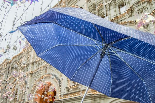 Close-up of an open blue umbrella with small polka dots, in the rain in the open air against the background of a city street.