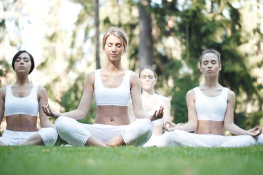 Women in white sportswear sitting in lotus position during group yoga training at park