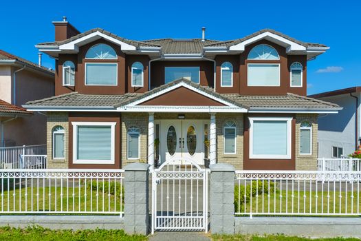 Big family house in suburban of Vancouver on blue sky background. Luxury residential house with iron fence and green lawn in front