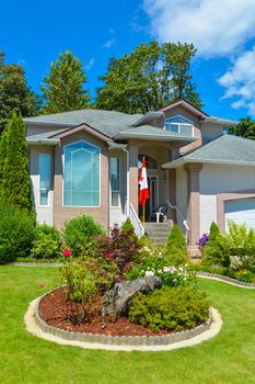 Residential house with Canadian flag at the entrance and nicely decorated front yard on blue sky background. Family house in British Columbia, Canada.