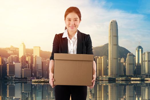 A young woman carrying a box wtih smile in cityscape background
