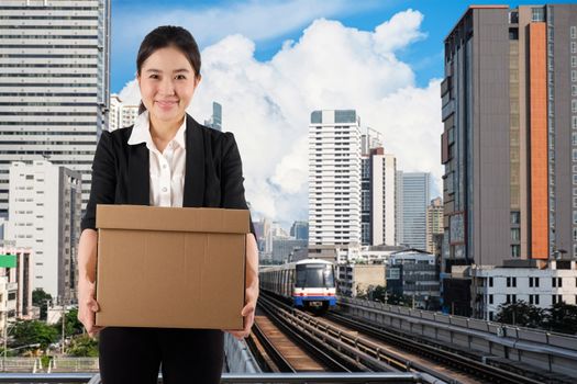 A young woman carrying a box wtih smile in city skytrain background