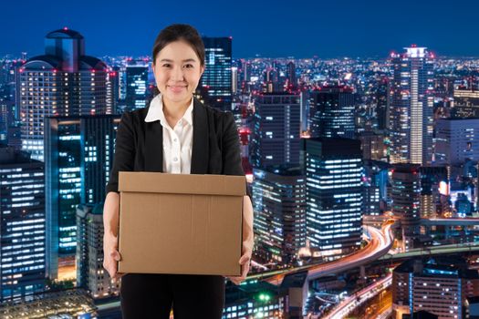 A young woman carrying a box wtih smile in night cityscape background
