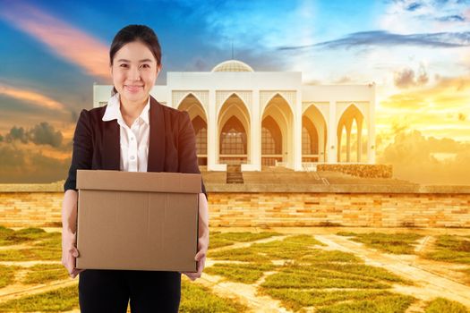 A young woman carrying a box wtih smiling on sunset in mosque background
