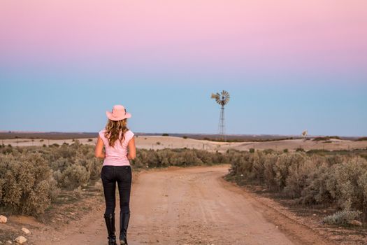 Woman walking along a dirt road of vast open spaces in outback NSW Australia  Windmill sits in a waterless landscape