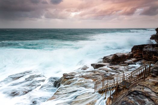 Large swells engulf the rock shelf right up the metal stairs, the resulting flows leaving a motion trail.  sunlight lights up the clouds in some soft hues.  Sydney