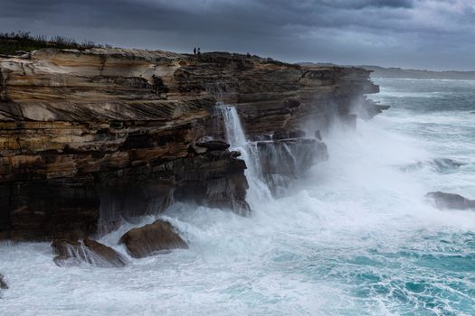 Large swells batter the cliffs of Sydney coast.  The larger swells smash against the cliff face throwing water upwards and then creating long cascading waterfalls of  around 20 metres height