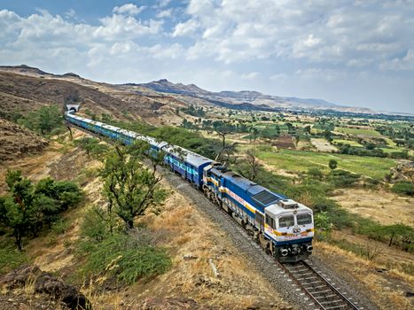 Shindawane, Pune, India:January 16th, 2016-A long train exits a tunnel on a clear background of blue sky with clouds.