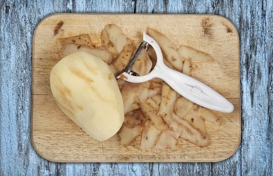 Raw potato peeled on a wooden chopping board with peeler cut out and isolated on a blue background