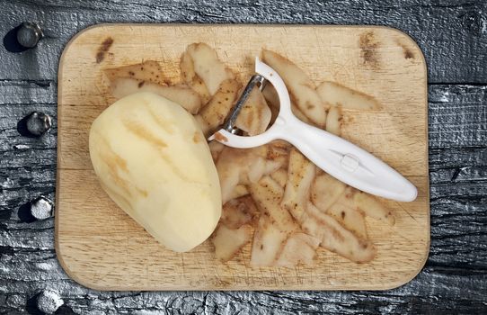 Raw potato peeled on a wooden chopping board with peeler cut out and isolated on a grey wood background