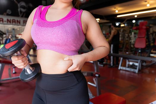 Fat woman holding dumbbell and excessive fat belly, overweight fatty belly at fitness gym. Diet lifestyle, weight loss, stomach muscle, healthy concept.