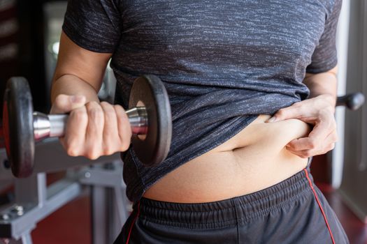 Fat man holding dumbbell and excessive fat belly, overweight fatty belly at fitness gym. Diet lifestyle, weight loss, stomach muscle, healthy concept.