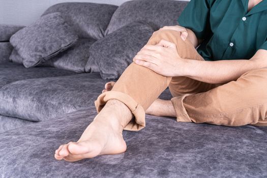 Young man suffering from knee pain while sitting on sofa at home. Healthcare medical or daily life concept.