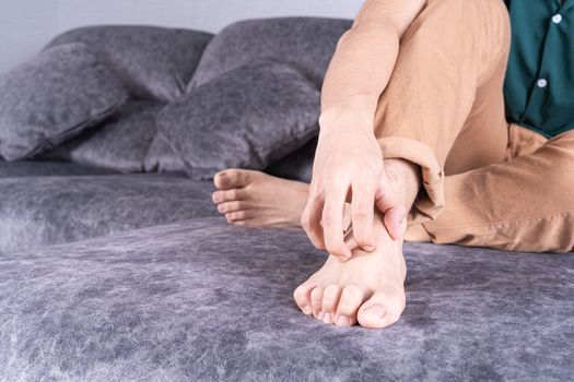 Young man scratching his foot while sitting on sofa at home. Healthcare medical or daily life concept.